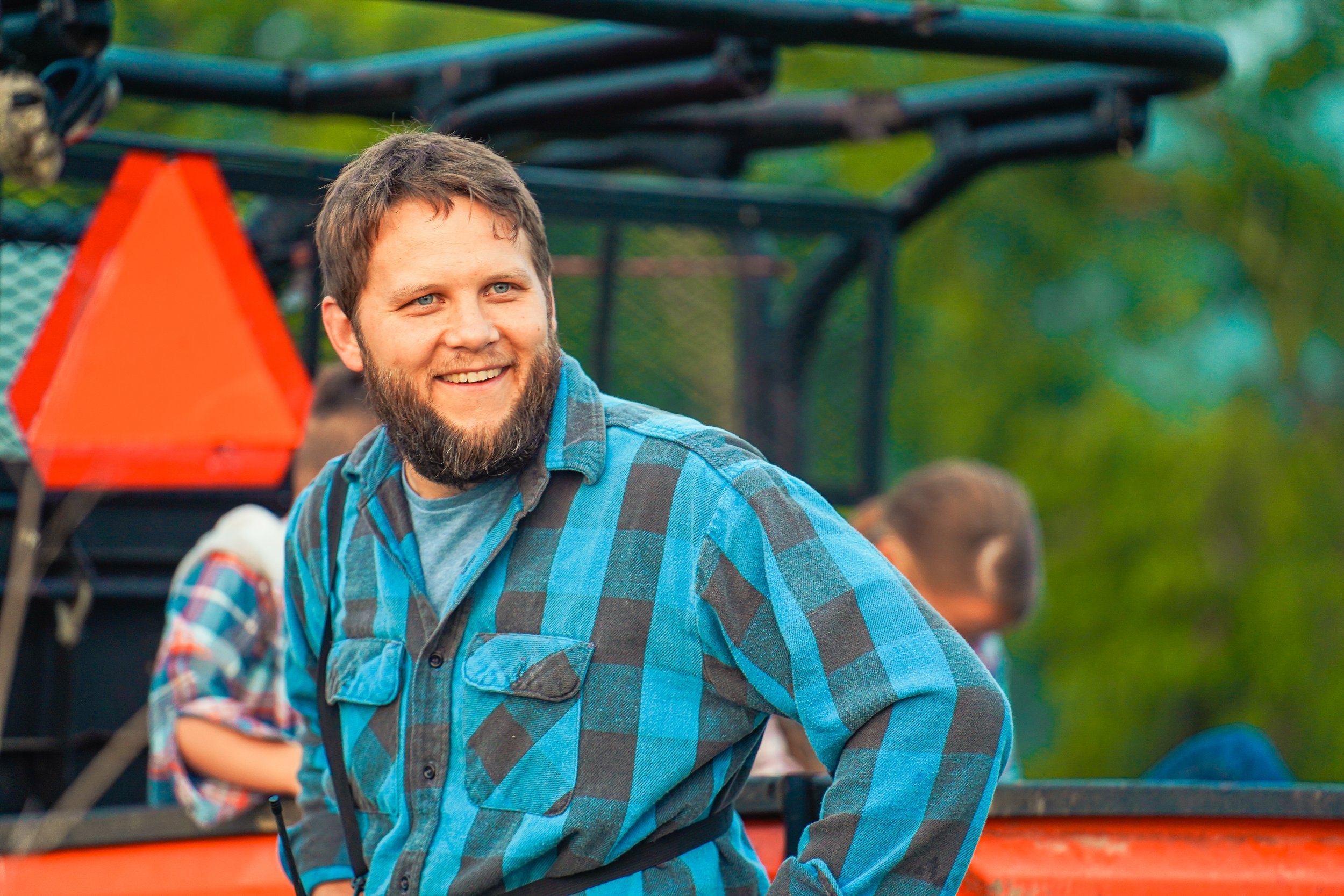 Smiling man in a blue and black checked shirt stands in front of a vehicle, surrounded by greenery, reflecting a relaxed outdoor atmosphere.
