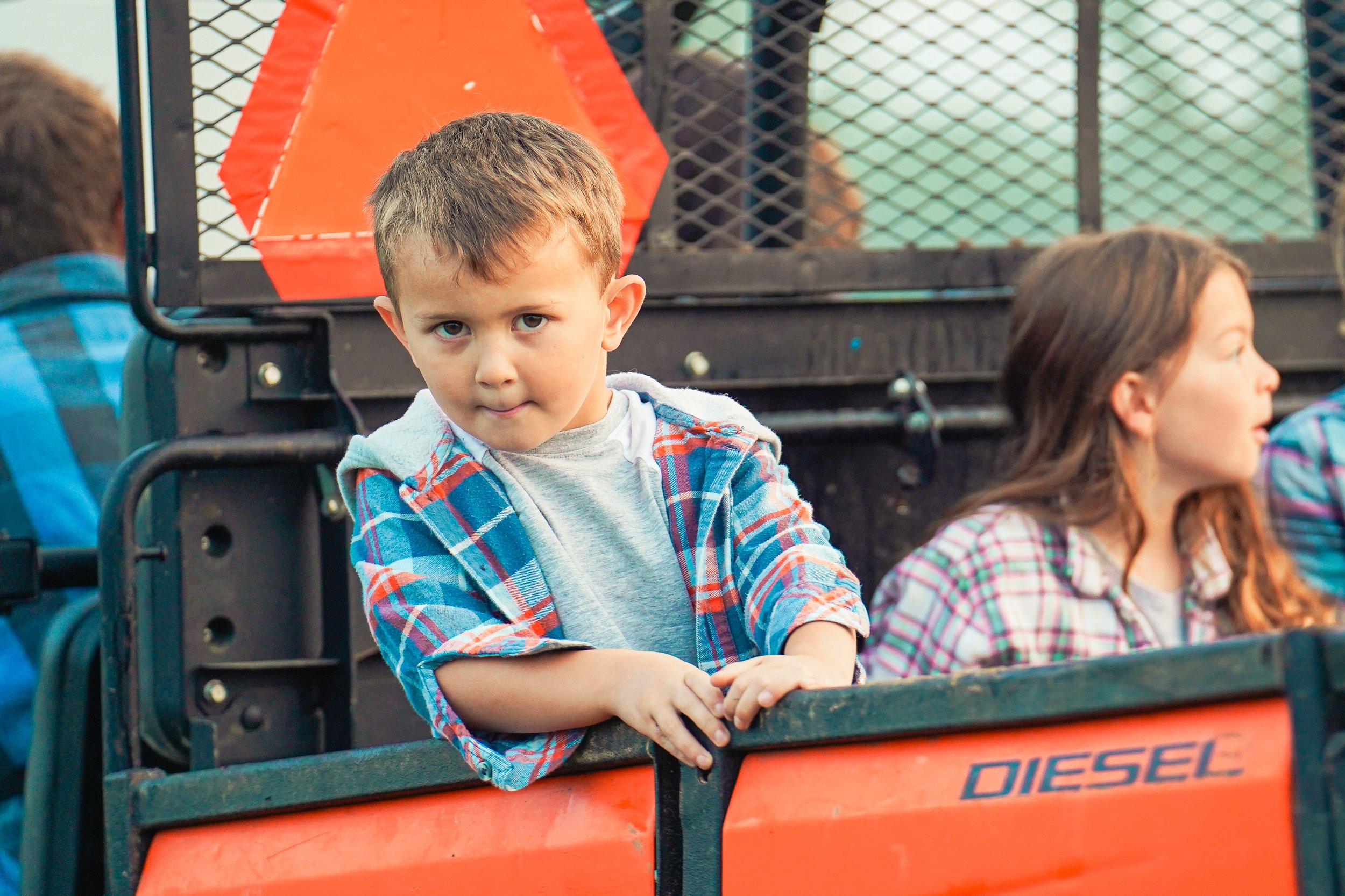 Young boy with short brown hair, wearing a gray shirt and a plaid flannel, leans against a truck while observing his surroundings, with a girl in the background also dressed in plaid. The scene takes place outdoors, highlighting a casual, playful atmosphere.