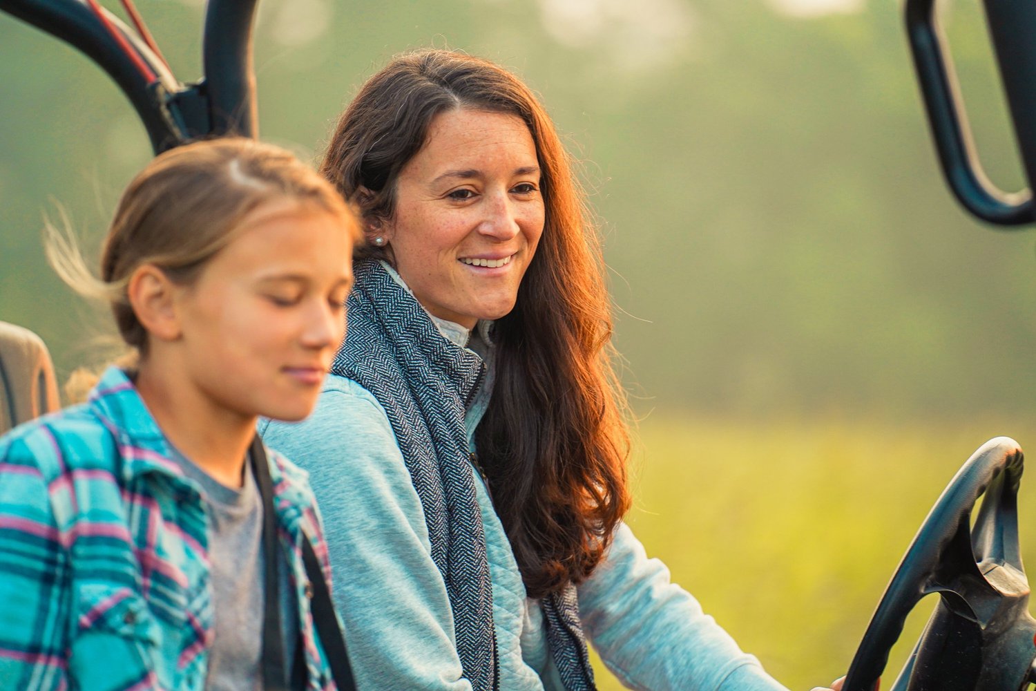 A woman and a girl enjoying a ride in a vehicle on a sunny day, surrounded by nature, showcasing a moment of connection and joy. The woman is smiling, while the girl appears thoughtful, both dressed casually in outdoor attire.