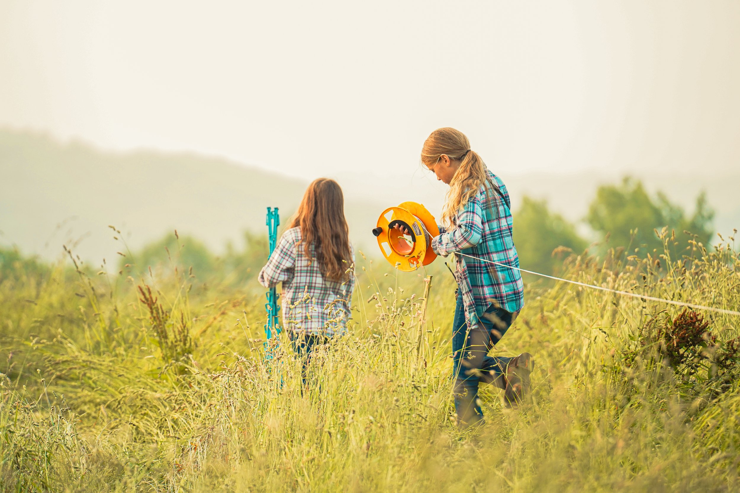 Two girls in plaid shirts walking through a grassy field, one holding a yellow measuring tape and the other carrying a blue tool, engaged in outdoor exploration or surveying.