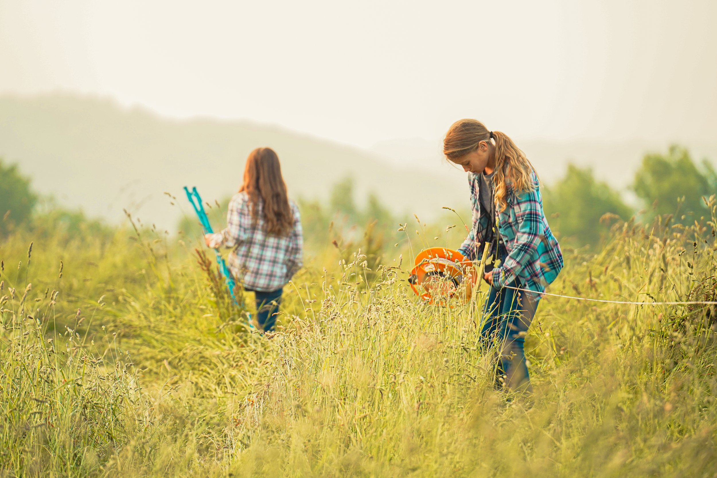 Two girls working in a lush, green field, one holding a measuring tape and the other using gardening tools, dressed in plaid shirts, showcasing outdoor activities and teamwork in nature.