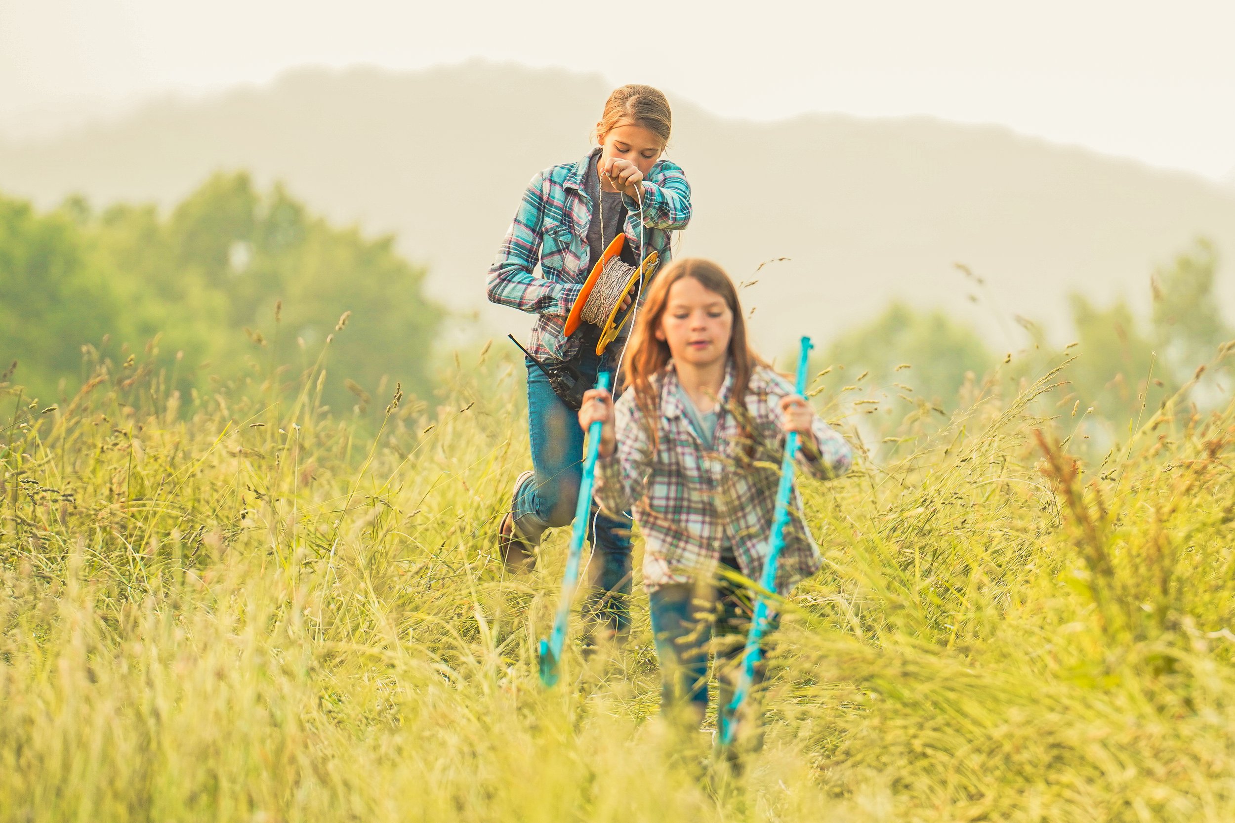 Two girls hiking through a grassy field, wearing plaid shirts and jeans, with one holding a fishing reel and the other carrying a blue hiking stick. The background features a soft, misty landscape, creating a serene outdoor atmosphere.