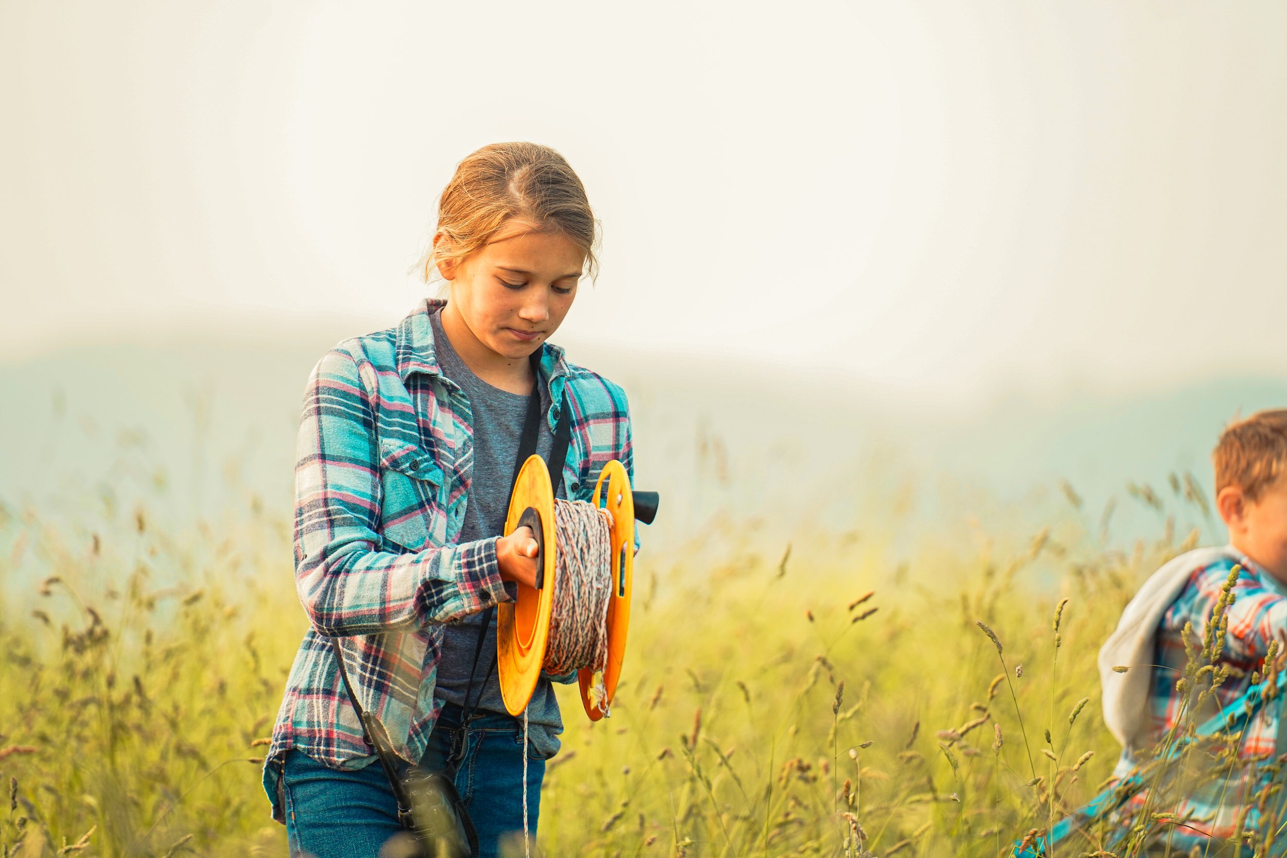 Young girl in a plaid shirt holding a spool of rope while standing in a grassy field, with a boy in the background. The scene captures outdoor activities and childhood exploration.