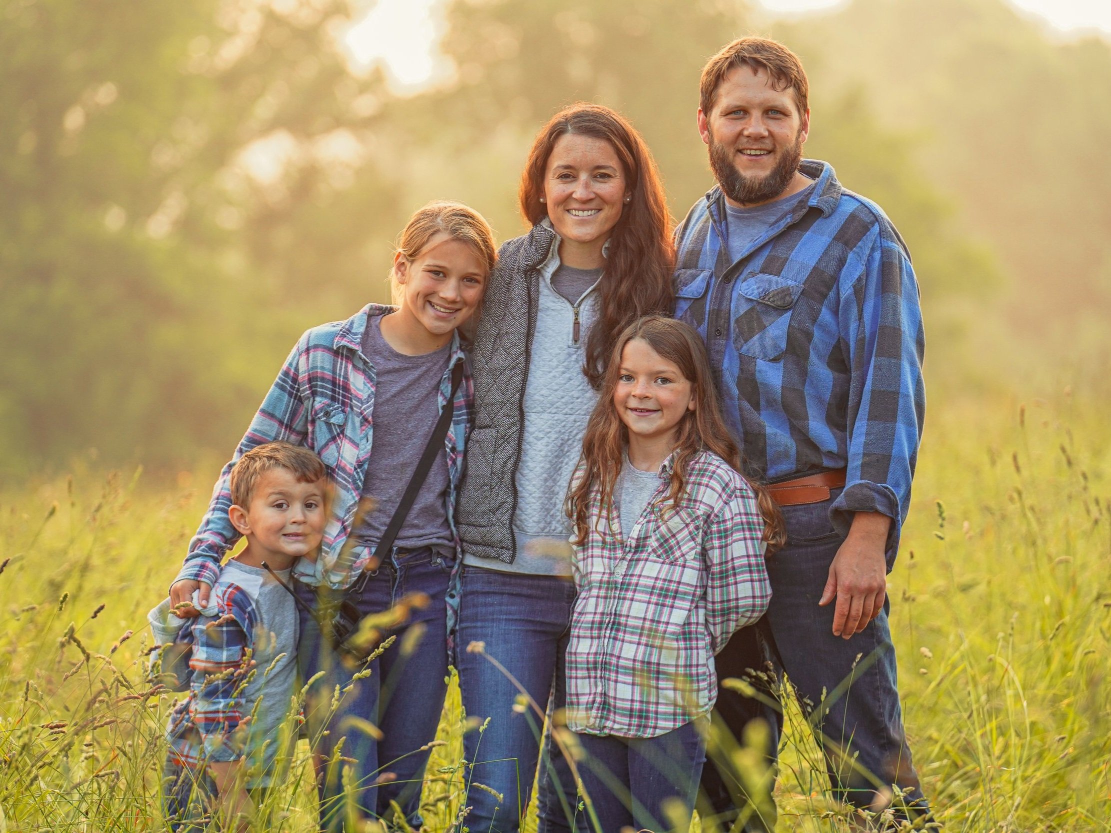 Family portrait of four smiling children and adults standing together in a sunlit field, surrounded by tall grass. The group is dressed in casual, plaid clothing, showcasing a warm, friendly atmosphere.
