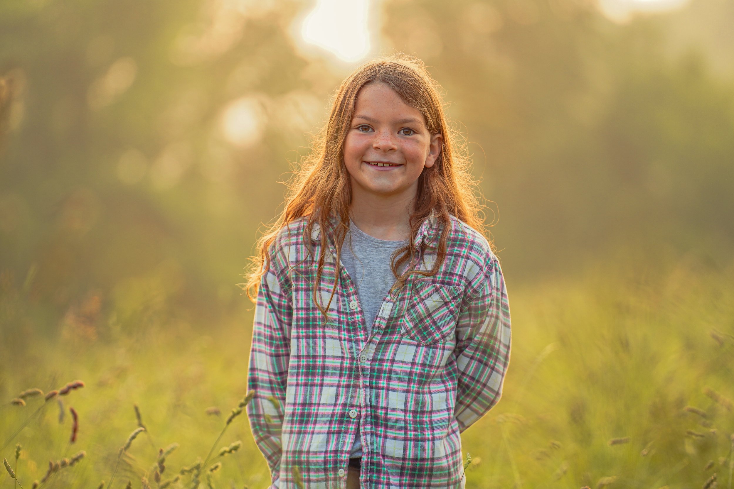Smiling child with long hair standing in a sunlit field, wearing a plaid shirt and gray t-shirt, surrounded by tall grass and a soft, glowing background.