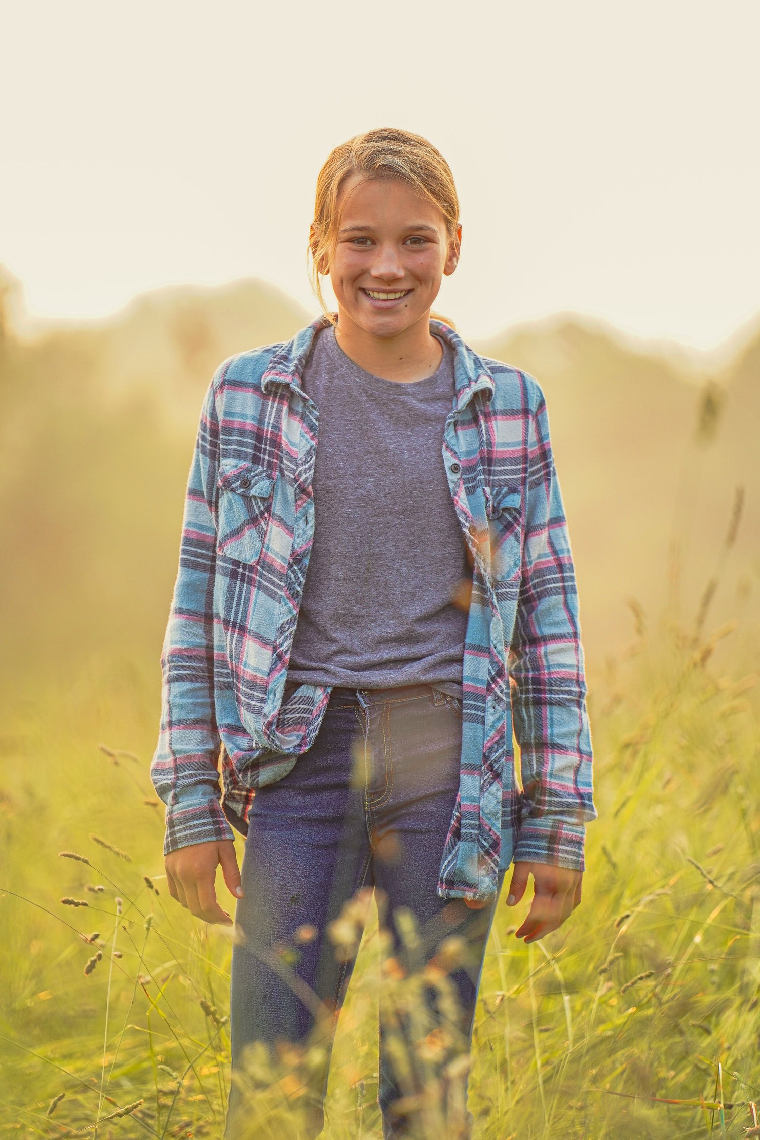 Smiling young girl wearing a plaid flannel shirt and jeans stands in a sunlit grassy field, surrounded by tall grass and soft light, capturing a joyful outdoor moment.