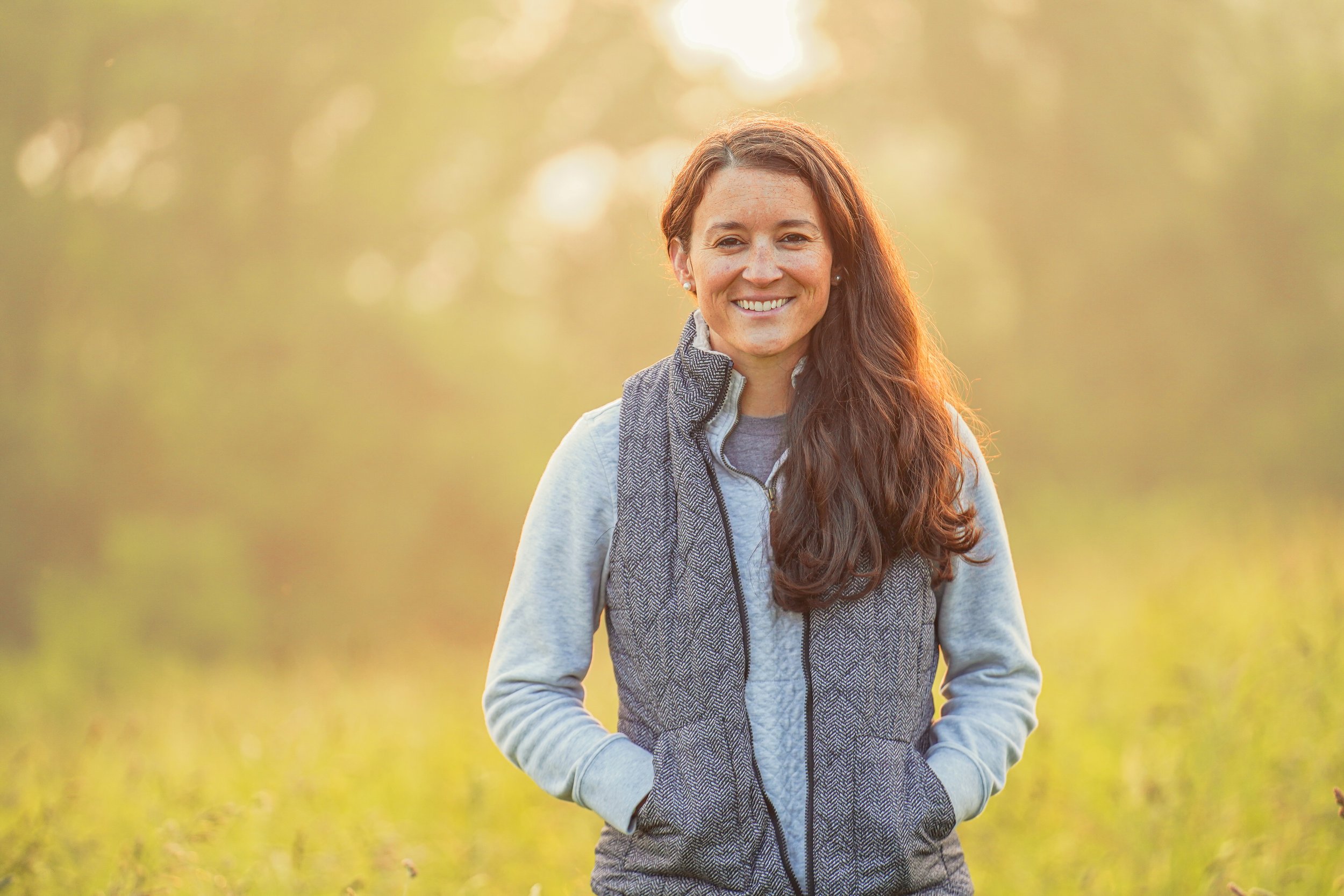 Smiling woman with long brown hair wearing a blue vest standing in a sunlit field, surrounded by greenery. The warm lighting creates a soft, inviting atmosphere, highlighting her cheerful expression.