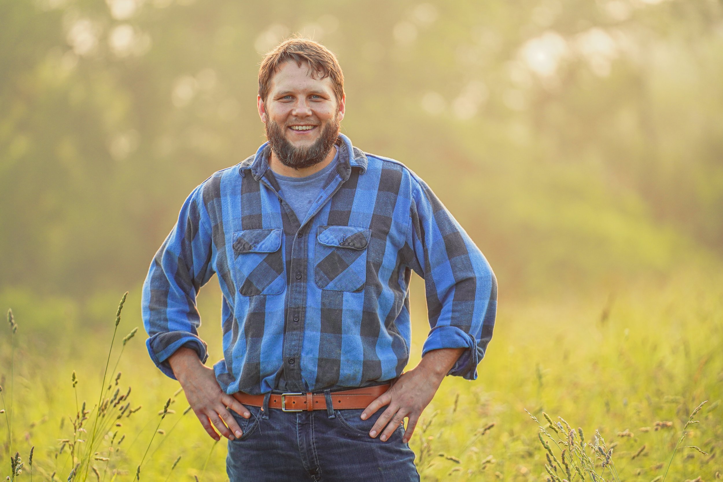 Smiling man in a blue and black checkered flannel shirt stands confidently in a sunlit field, surrounded by tall grass and a blurred green background.